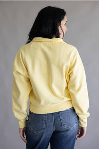 A woman viewed from behind wearing a pale yellow cropped sweatshirt and blue jeans against a plain gray studio background.