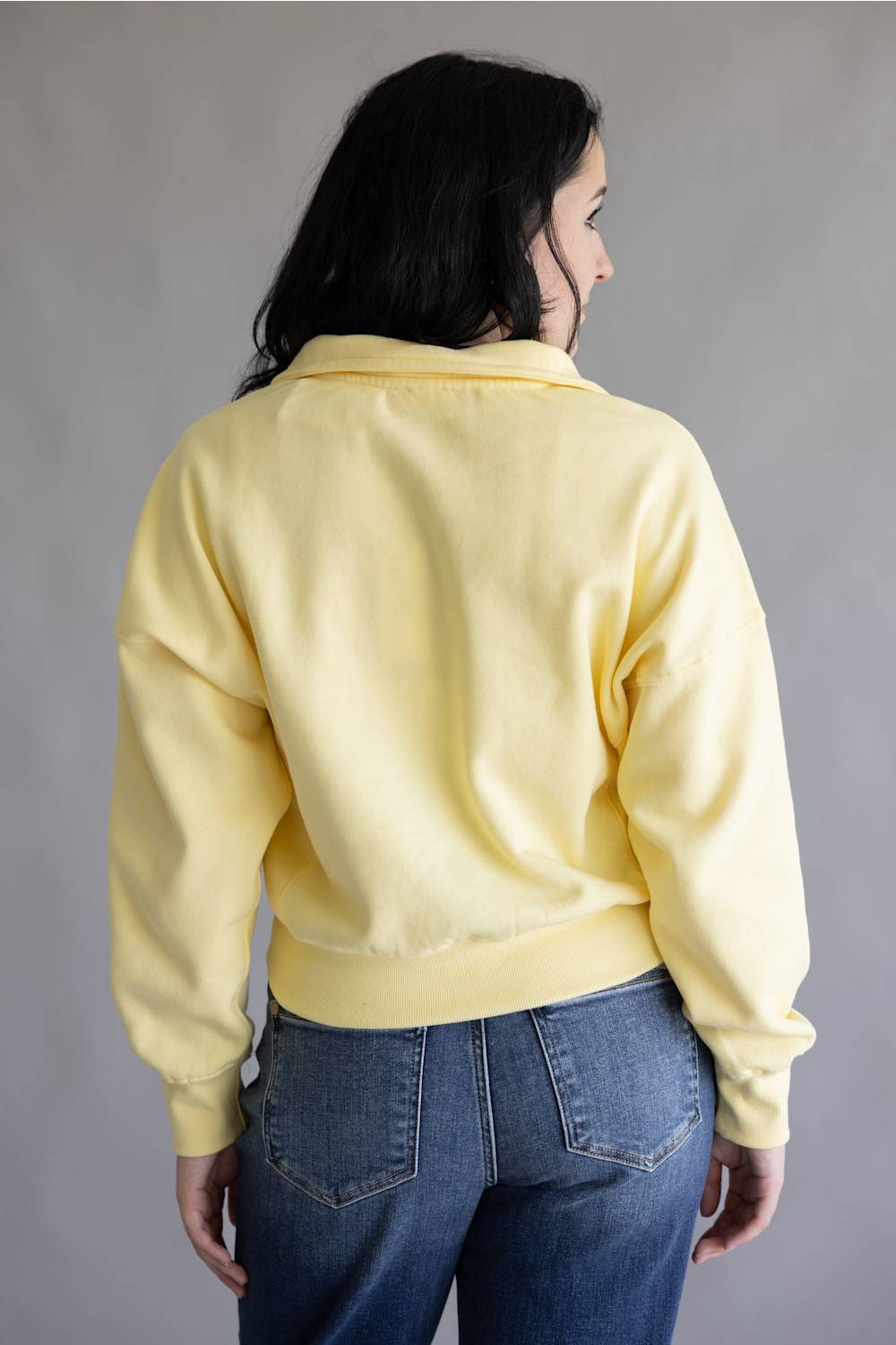 A woman viewed from behind wearing a pale yellow cropped sweatshirt and blue jeans against a plain gray studio background.