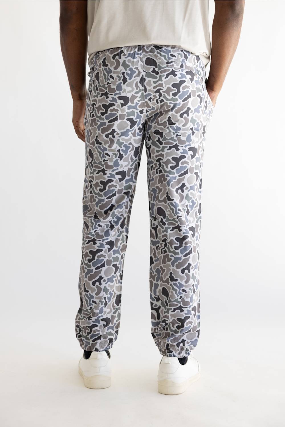 A person shown from the back wearing camouflage print pants with a gray, white, and black camo pattern, paired with a light beige t-shirt and white sneakers, against a plain white studio background.