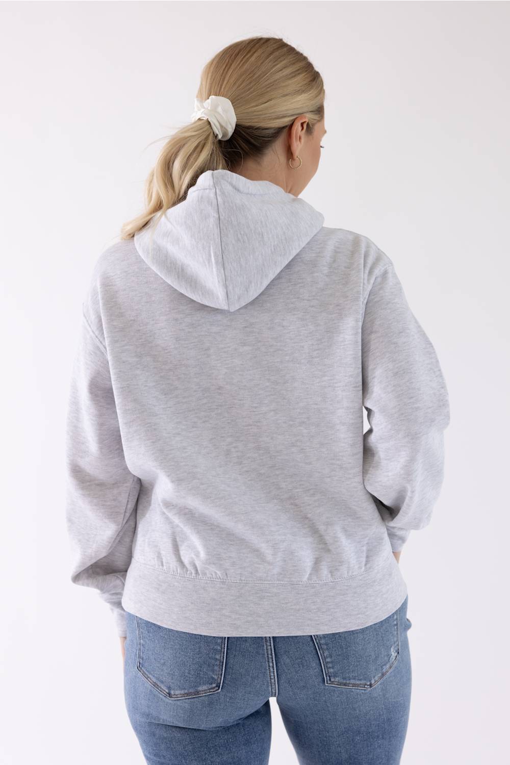 Back view of a blonde woman wearing an ice grey Reflex fleece pullover hoodie, blue jeans, and a white hair tie.