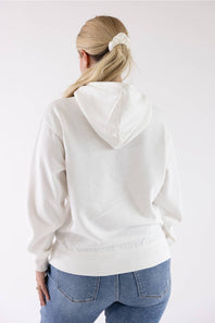 Back view of a woman with blonde hair tied back wearing an ivory Reflex fleece pullover hoodie and blue jeans.
