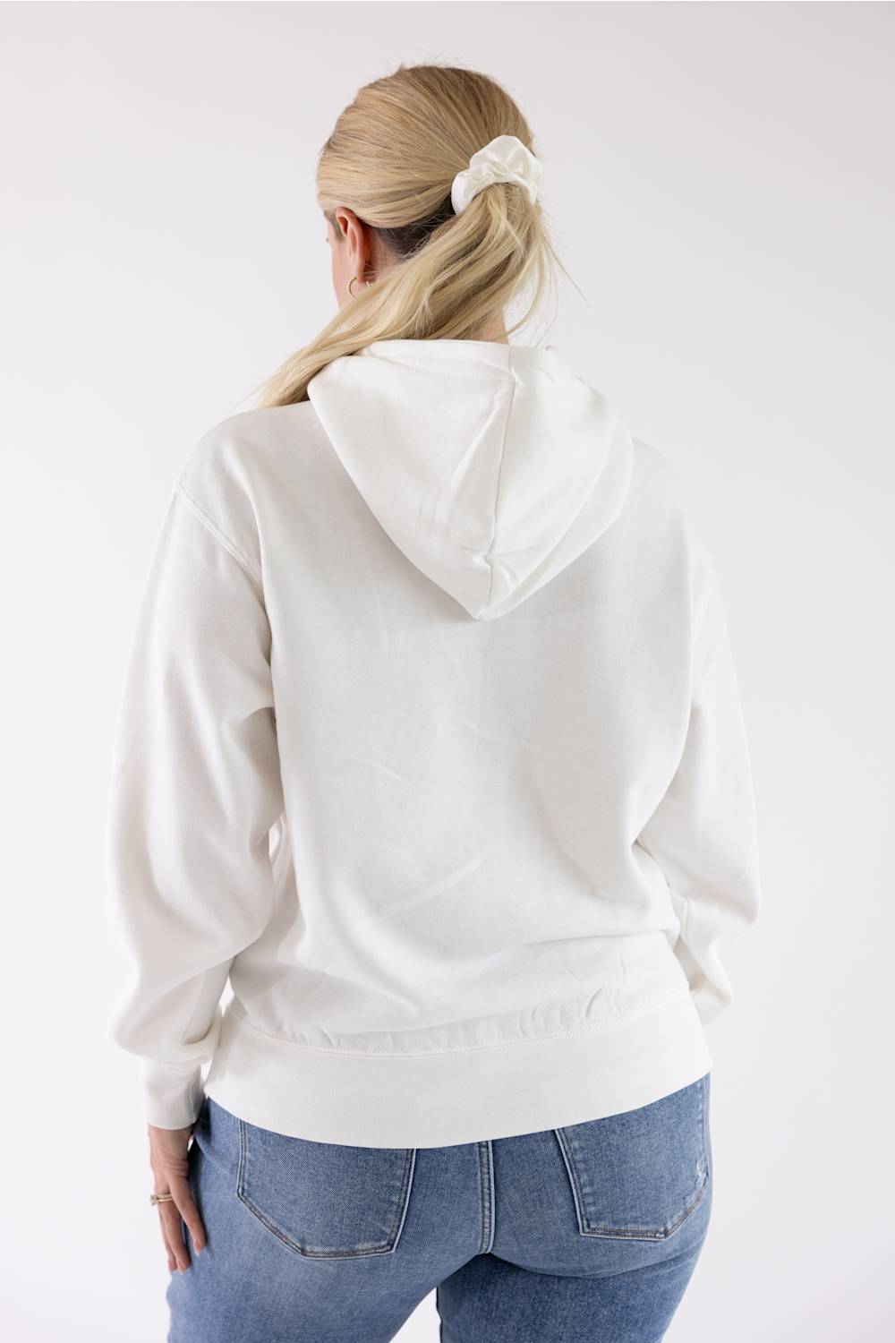 Back view of a woman with blonde hair tied back wearing an ivory Reflex fleece pullover hoodie and blue jeans.