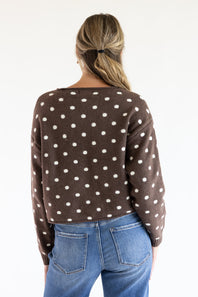 Back view of a model wearing the Just Polly dark brown long-sleeve knit sweater featuring an ivory polka dot pattern, a slightly rolled boat neckline, and ribbed cuffs, styled with blue jeans against a plain white background.