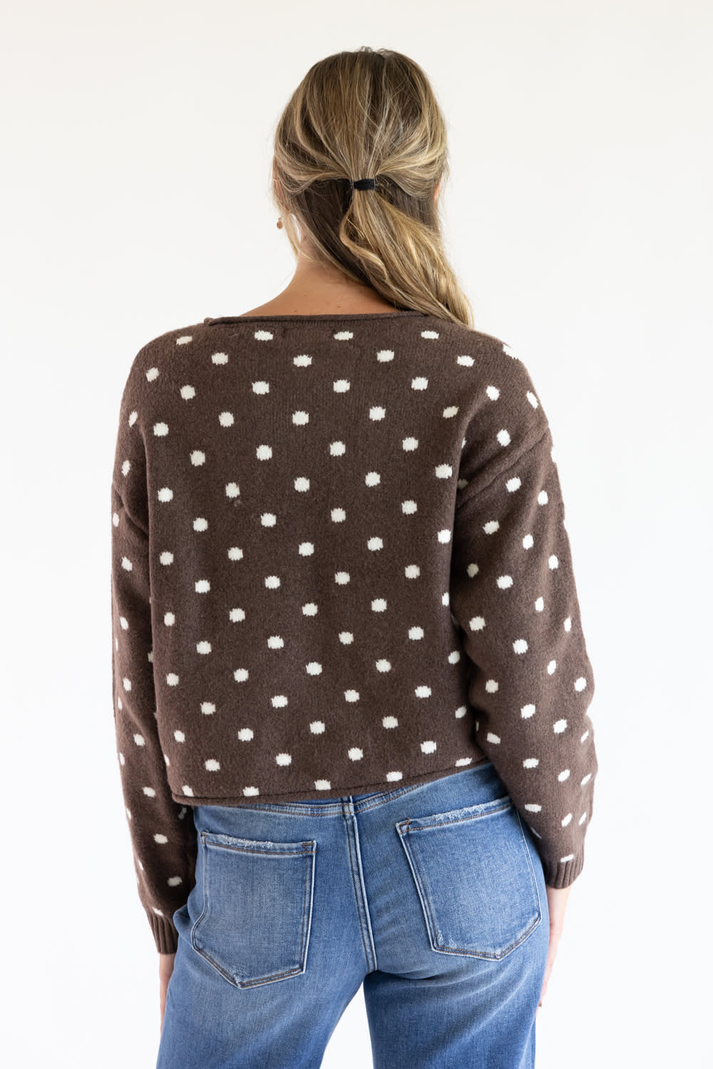 Back view of a model wearing the Just Polly dark brown long-sleeve knit sweater featuring an ivory polka dot pattern, a slightly rolled boat neckline, and ribbed cuffs, styled with blue jeans against a plain white background.