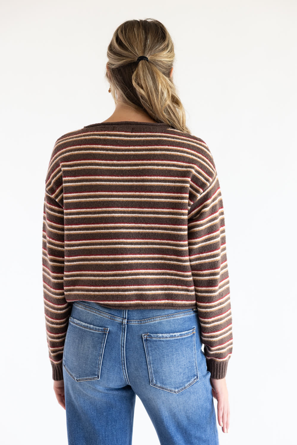 Back view of a model wearing the Just Polly long-sleeve knit sweater featuring multi-color horizontal stripes in brown, red, and tan, styled with denim jeans against a plain white background.