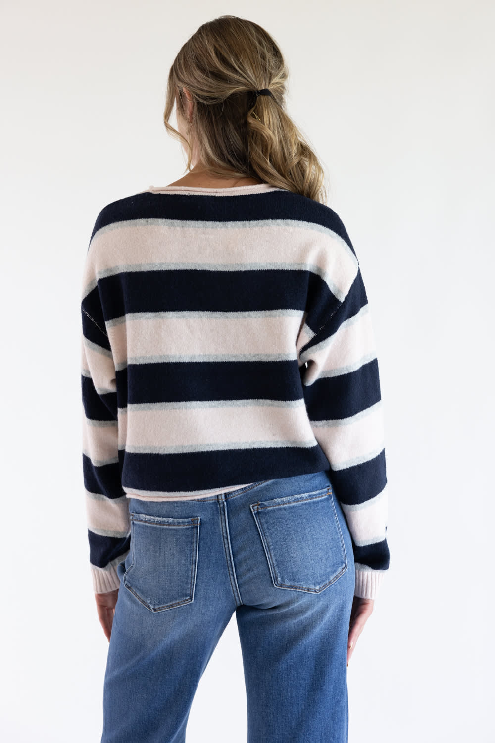 Back view of a model wearing the Just Polly long-sleeve knit sweater featuring wide pink and navy horizontal stripes with thin heather grey accents, styled with blue jeans against a plain background.