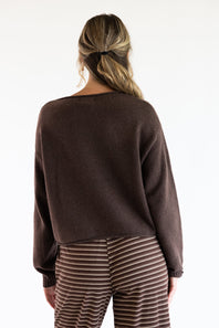 Back view of a model wearing the Just Polly dark brown knit boat neck sweater featuring long sleeves, a plain knit back, and a straight hemline, styled with brown and white striped trousers.