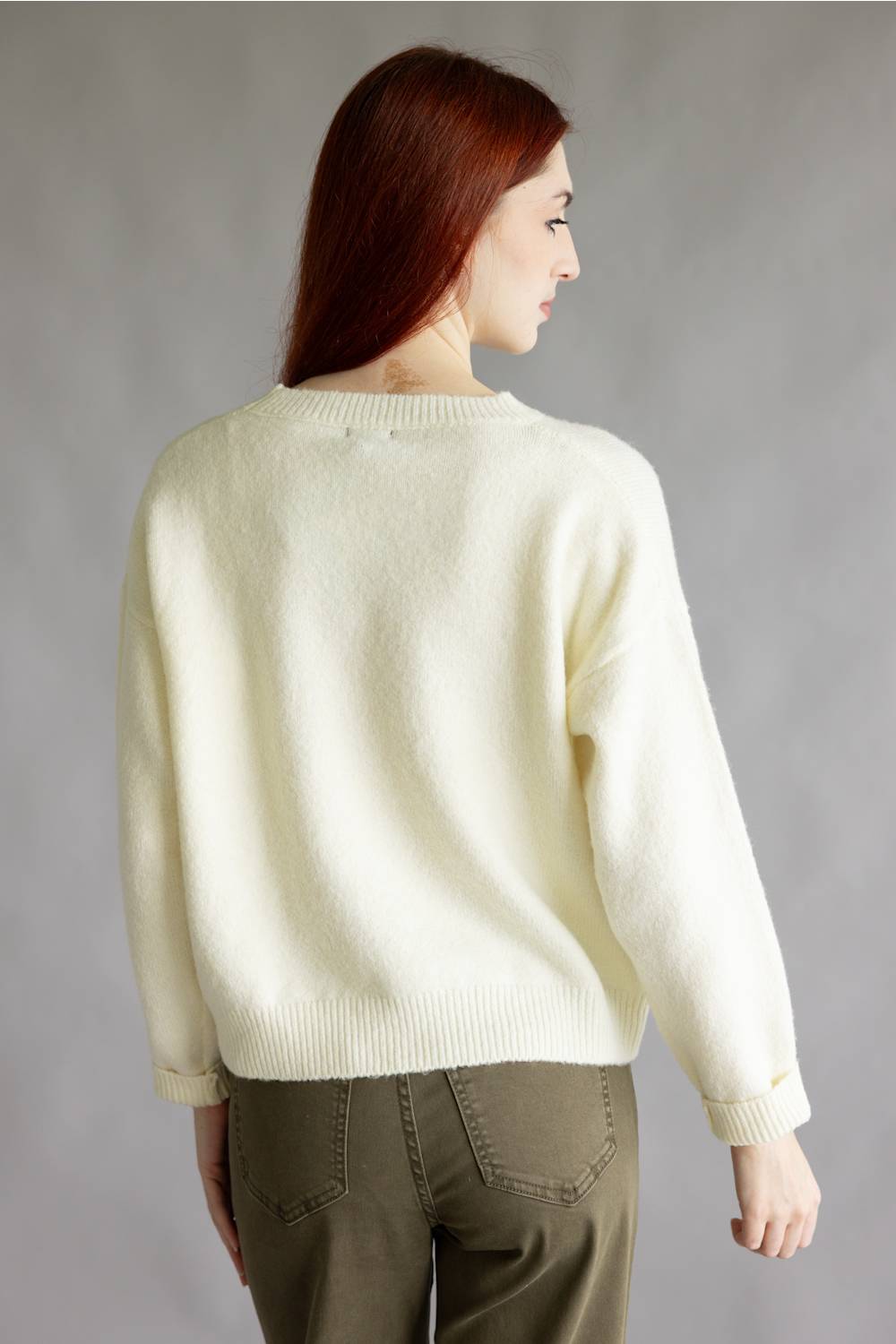 A woman with red hair wearing a cream-colored knit sweater with a detailed duck motif on the front, paired with olive green pants, standing against a neutral gray studio background.
