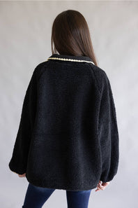 A woman viewed from behind wearing a black sherpa fleece jacket with a cream-colored beaded collar, paired with blue jeans, against a plain light gray background.
