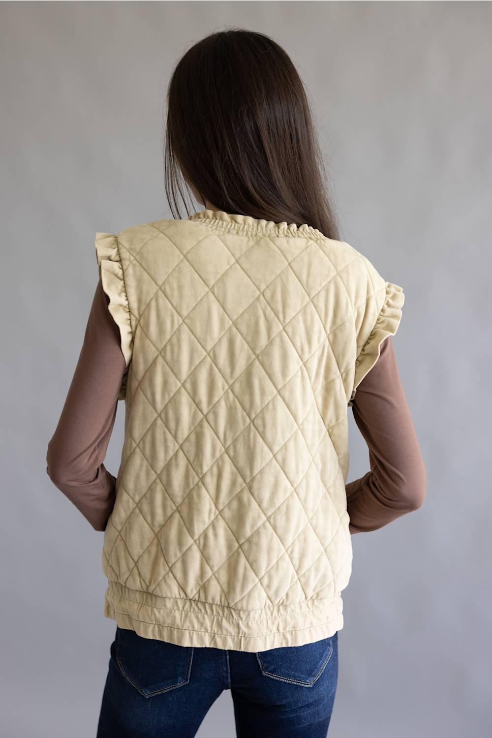 A woman viewed from behind wearing a cream-colored diamond-quilted sleeveless vest over a brown long-sleeve top and blue jeans, set against a plain gray studio background.