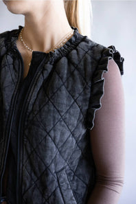 Close-up of a person wearing a black quilted sleeveless vest with ruffled shoulder details over a sheer nude long-sleeve top.