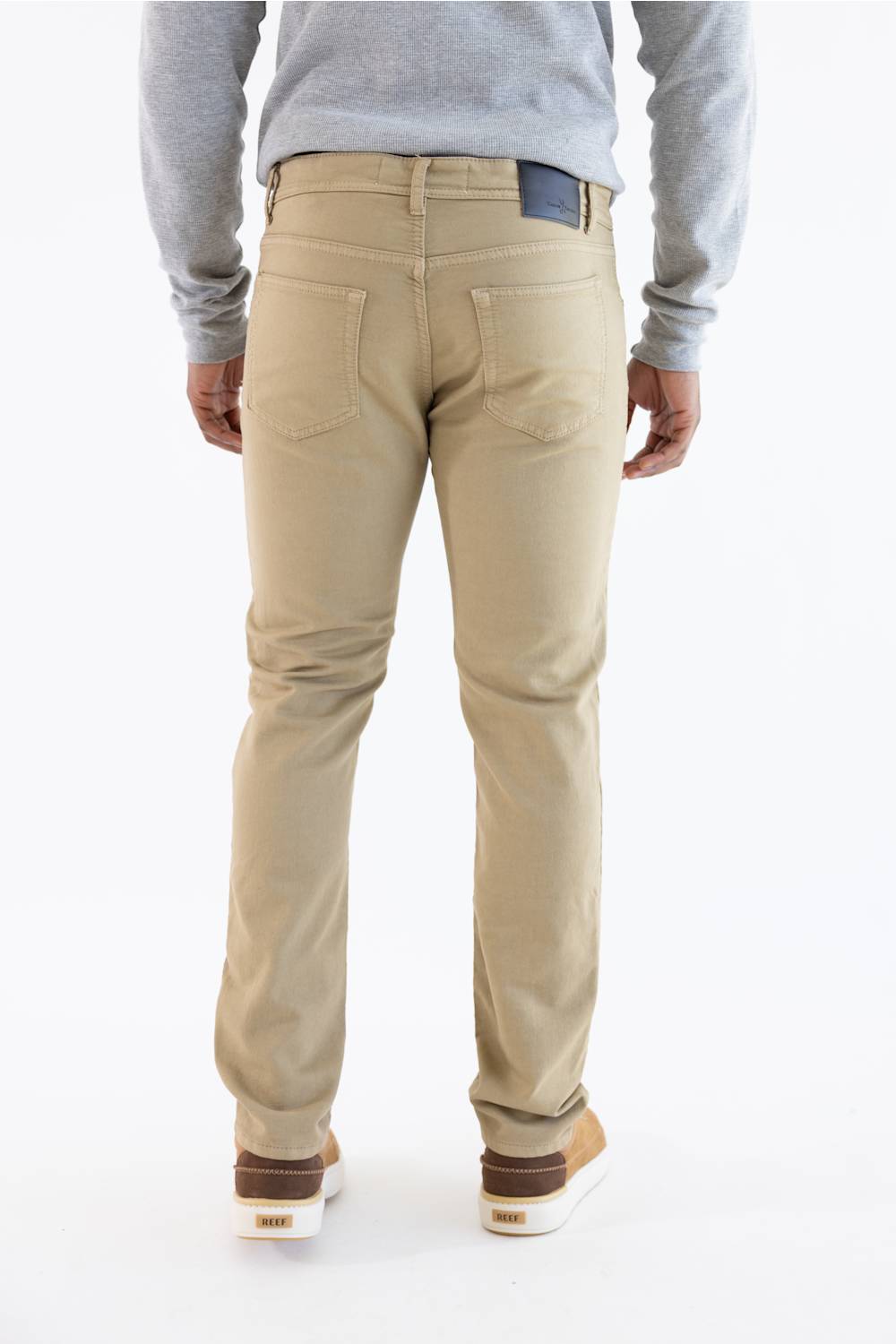 A person shown from the back wearing beige slim-fit chino pants, a light gray long-sleeve shirt, and brown and white sneakers against a plain white studio background.