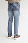 Rear view of a person wearing light-wash blue denim jeans with brown shoes, standing against a plain light gray studio background.