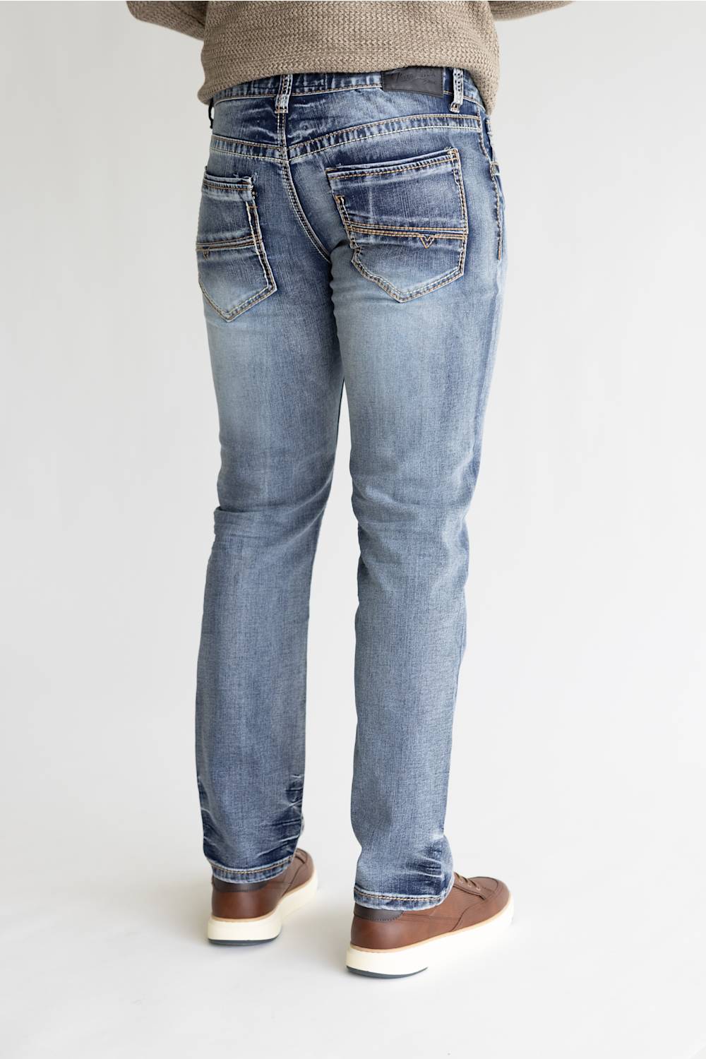 Rear view of a person wearing light-wash blue denim jeans with brown shoes, standing against a plain light gray studio background.