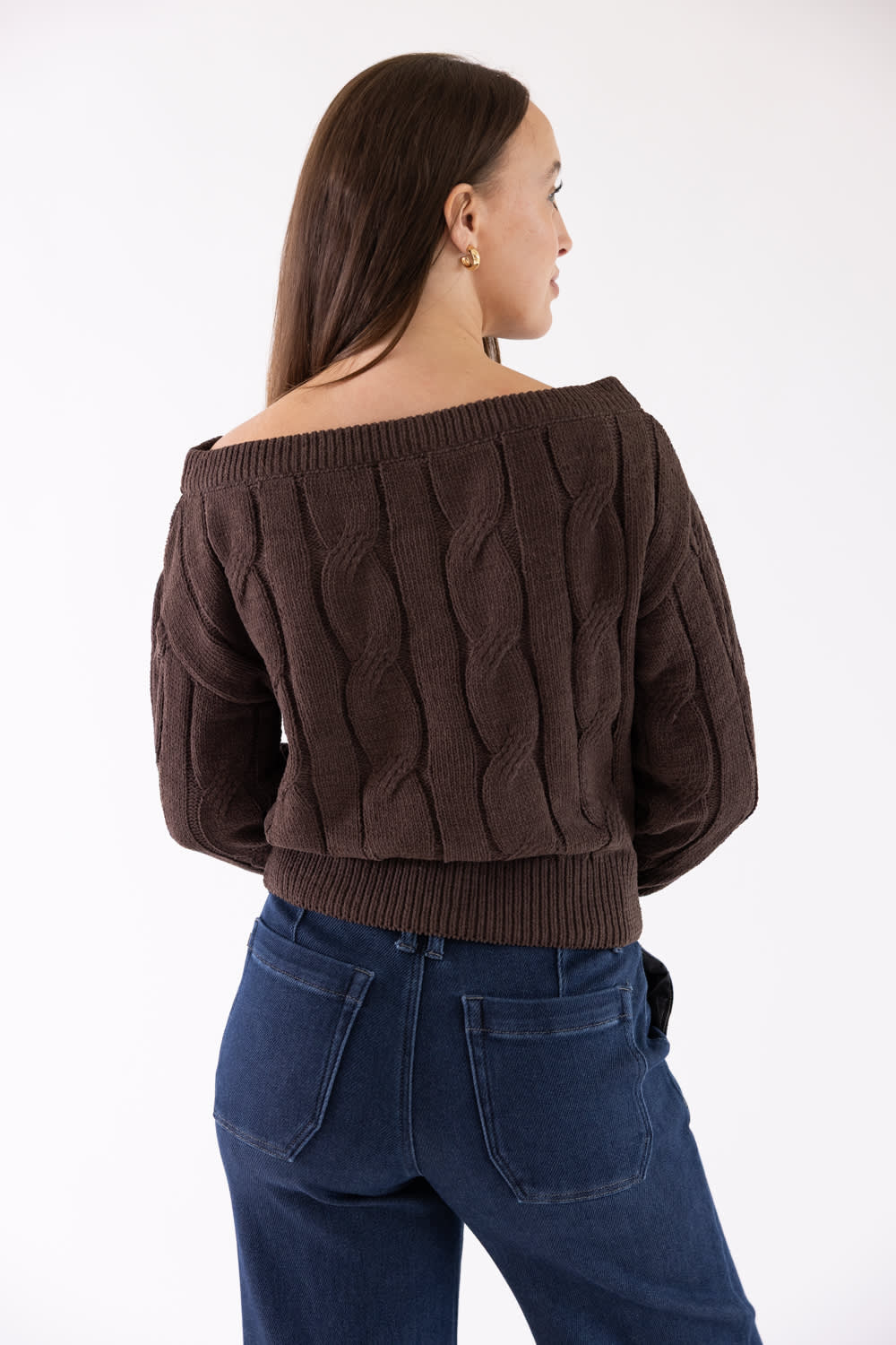 A woman shown from the back wearing a dark brown off-the-shoulder cable-knit cropped sweater and dark blue jeans, photographed against a plain white studio background.