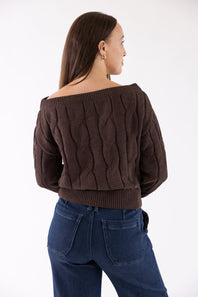 A woman shown from the back wearing a dark brown off-the-shoulder cable-knit cropped sweater and dark blue jeans, photographed against a plain white studio background.