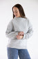 A woman wearing a light gray heathered knit sweater with a mock neck and ribbed cuffs, paired with blue jeans with small white dots, against a plain white studio background.