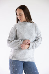 A woman wearing a light gray heathered knit sweater with a mock neck and ribbed cuffs, paired with blue jeans with small white dots, against a plain white studio background.