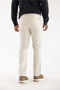A man viewed from the back wearing light beige chino pants, a dark navy long-sleeve shirt, and brown shoes, against a plain white studio background.