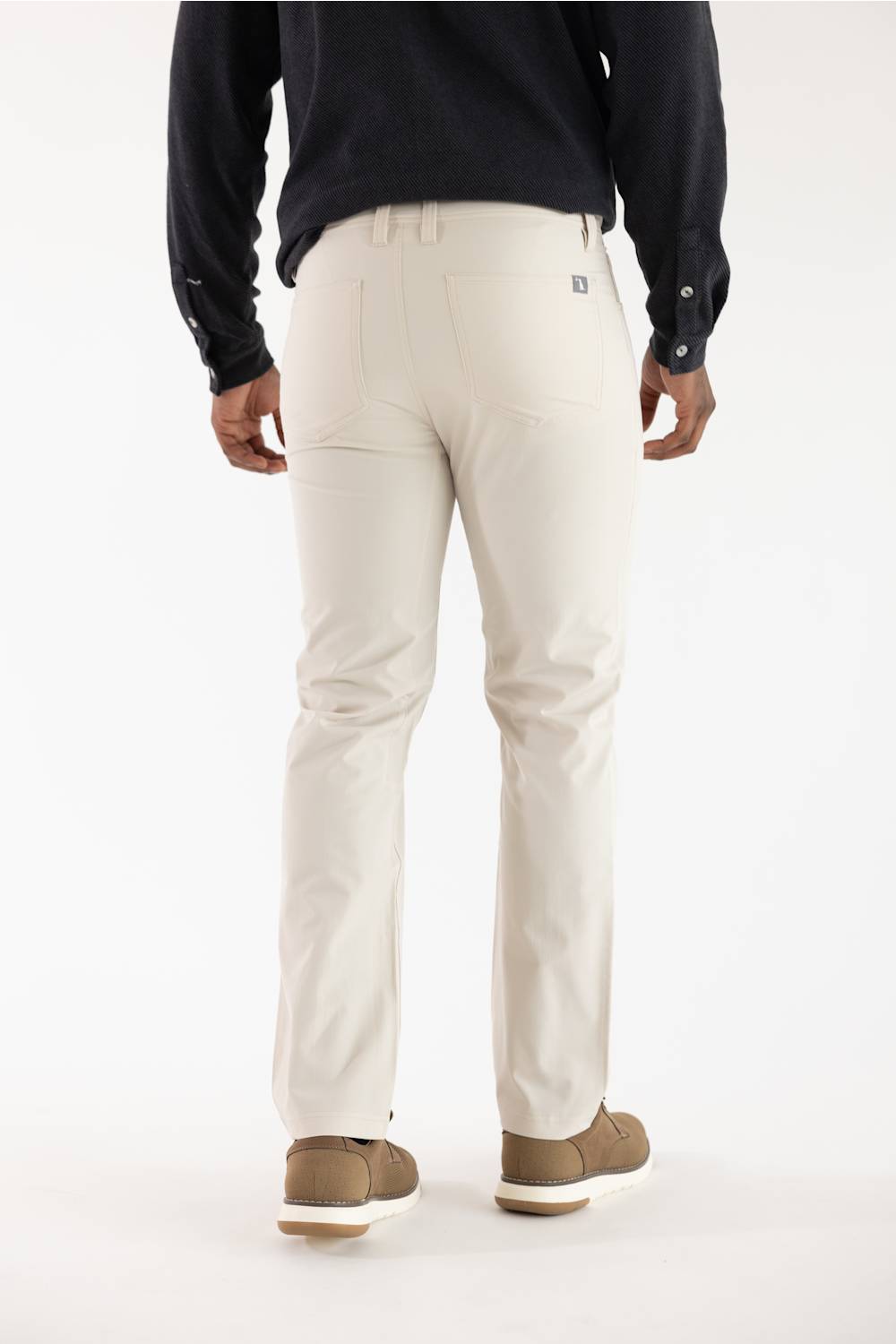 A man viewed from the back wearing light beige chino pants, a dark navy long-sleeve shirt, and brown shoes, against a plain white studio background.