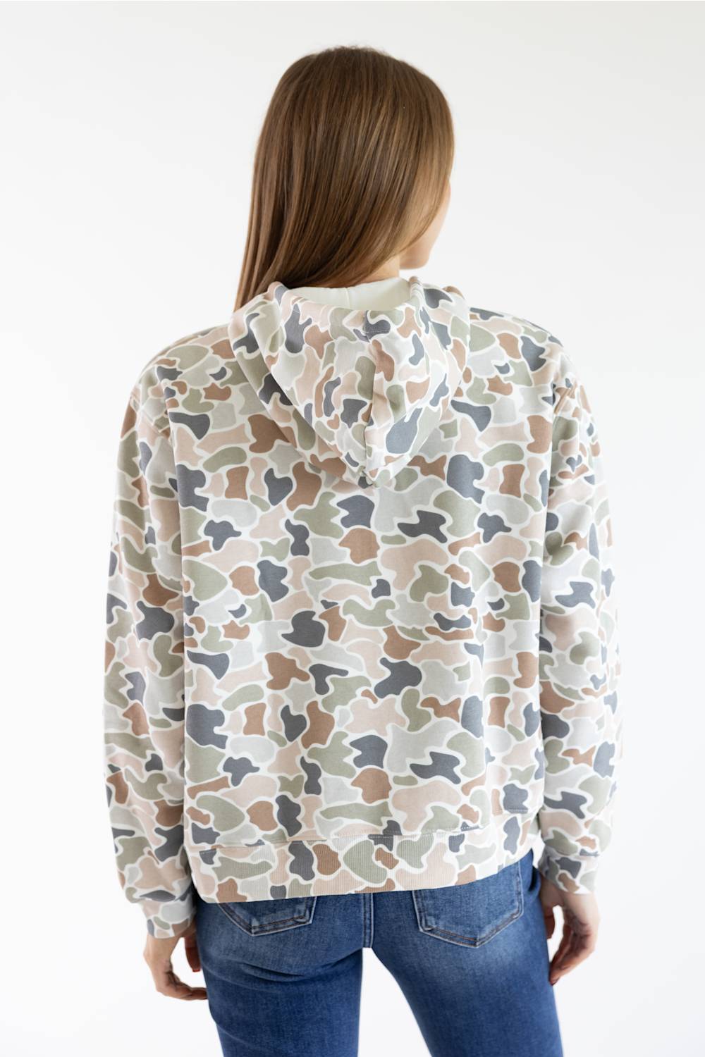 A woman viewed from the back wearing a camouflage hoodie with a beige, brown, gray, and green pattern, paired with blue jeans, against a plain white background.