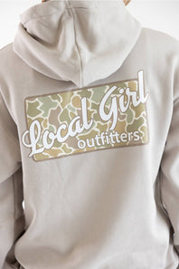 Close-up of the back of a light beige hoodie featuring a large graphic patch with a camouflage pattern and the text 'Local Girl outfitters' in white script.