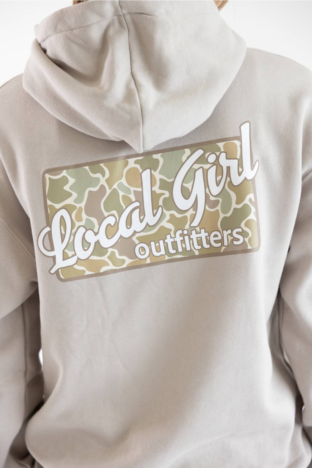 Close-up of the back of a light beige hoodie featuring a large graphic patch with a camouflage pattern and the text 'Local Girl outfitters' in white script.