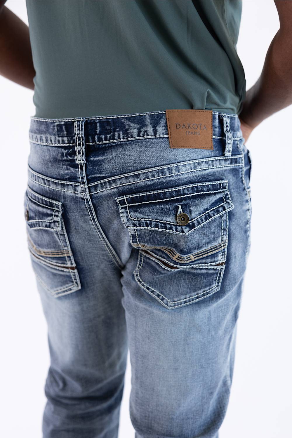 Close-up back view of a person wearing light-wash blue denim jeans with a tan leather DAKOT brand patch on the waistband, paired with a muted green top against a white background.