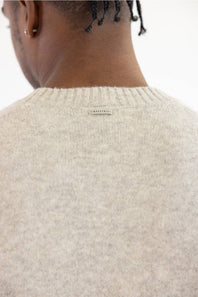 Close-up back view of a person wearing a cream-colored knit sweater, showing the ribbed crew neck and a small woven label at the center.