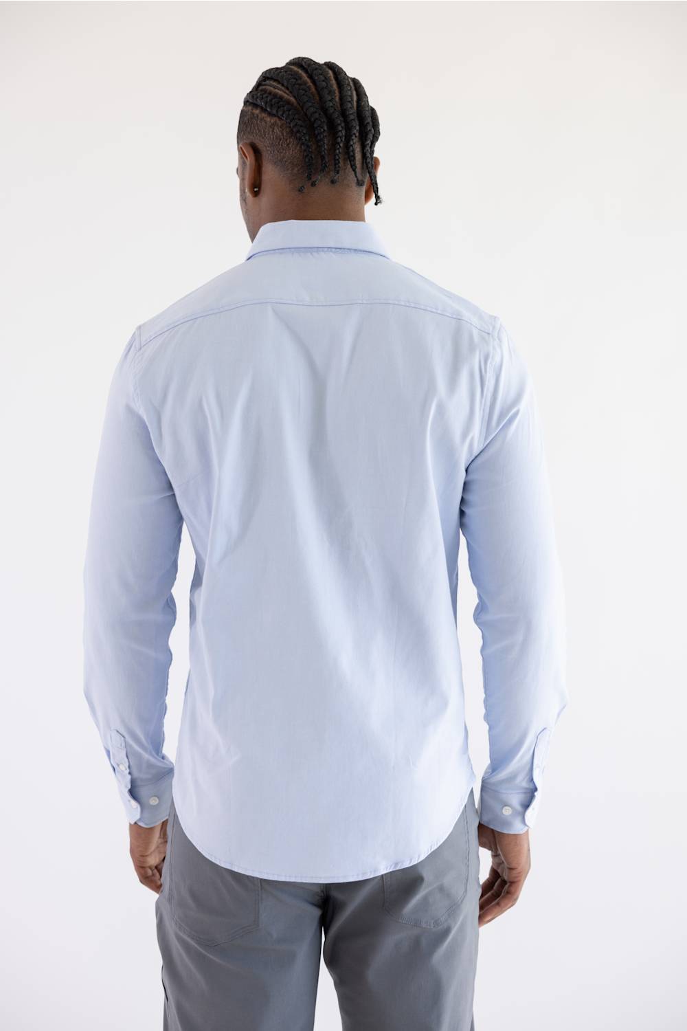 A man viewed from behind wearing a light blue long-sleeve button-down shirt and gray trousers, standing against a plain white studio background.