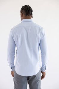 A man viewed from behind wearing a light blue long-sleeve button-down shirt and gray trousers, standing against a plain white studio background.