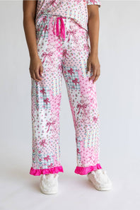 A person wearing pink and white patterned pajama pants with a ruffled hem, shown from the waist down against a light gray studio background. The pants have a patchwork print with bows, polka dots, gingham, and floral motifs, paired with white fluffy slippers.