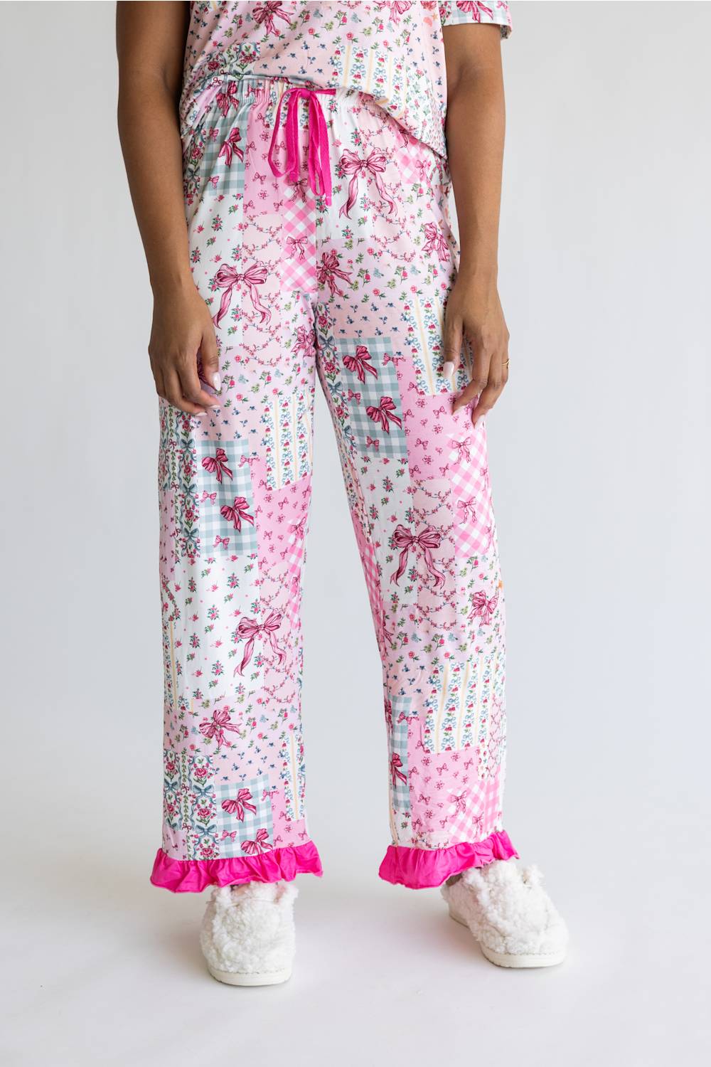 A person wearing pink and white patterned pajama pants with a ruffled hem, shown from the waist down against a light gray studio background. The pants have a patchwork print with bows, polka dots, gingham, and floral motifs, paired with white fluffy slippers.