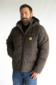 A man wearing a brown Carhartt puffer jacket with a hood, paired with a black shirt and blue jeans, standing against a light gray background.