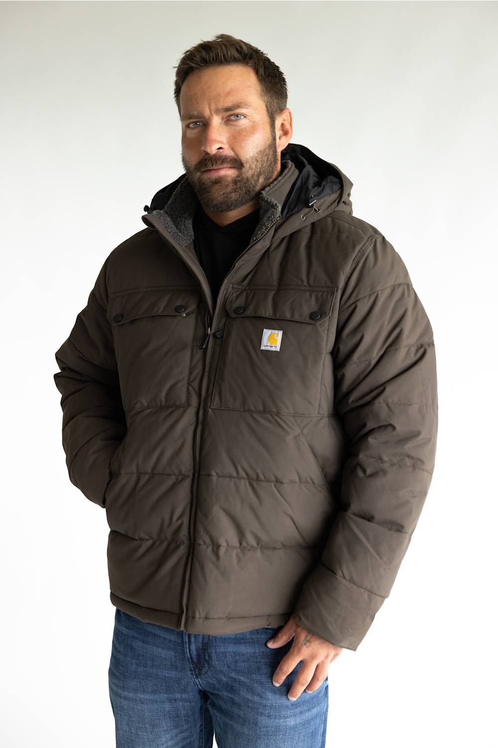 A man wearing a brown Carhartt puffer jacket with a hood, paired with a black shirt and blue jeans, standing against a light gray background.