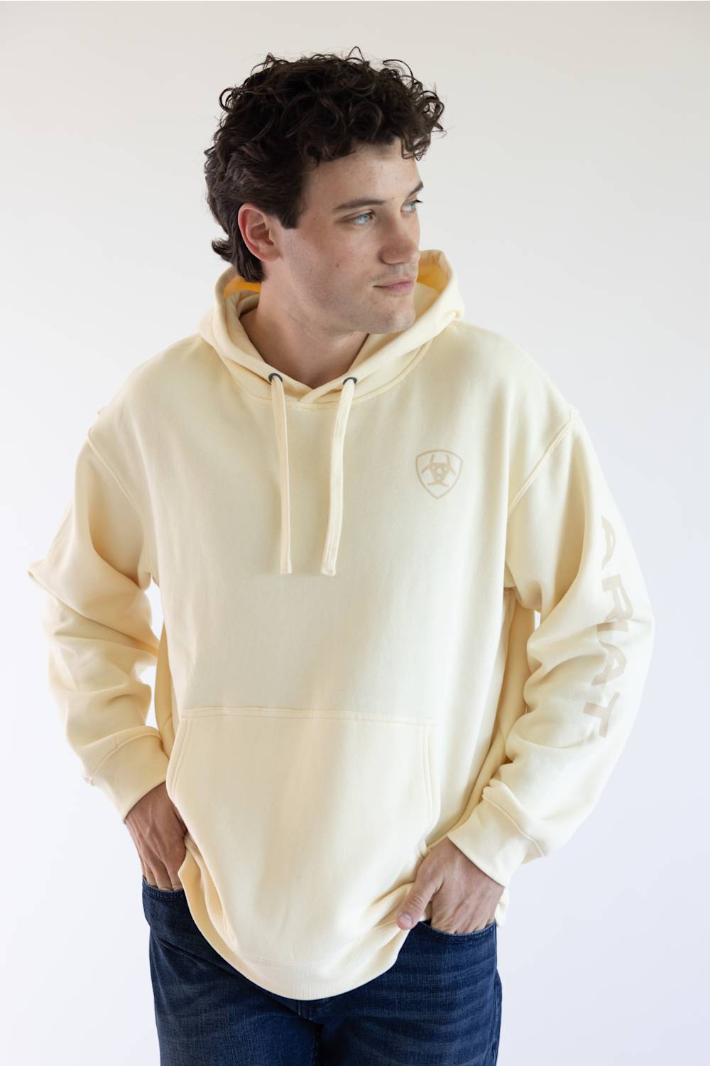 A man wearing a cream-colored pullover hoodie with a small shield logo on the left chest, paired with dark blue jeans, photographed against a plain white studio background.
