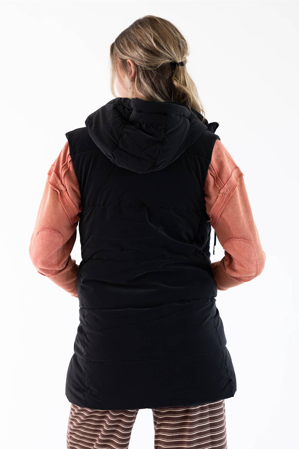 A woman viewed from behind wearing a black hooded puffer vest over a peach-pink long-sleeve top and brown-and-white striped pants, against a plain white studio background.