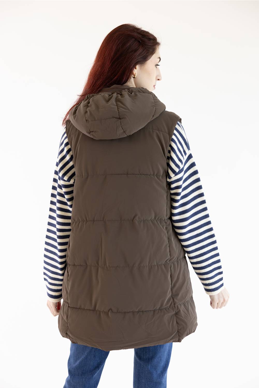 A woman viewed from the back wearing a brown hooded puffer vest over a navy and white striped long-sleeve shirt, paired with blue jeans, against a plain white background.
