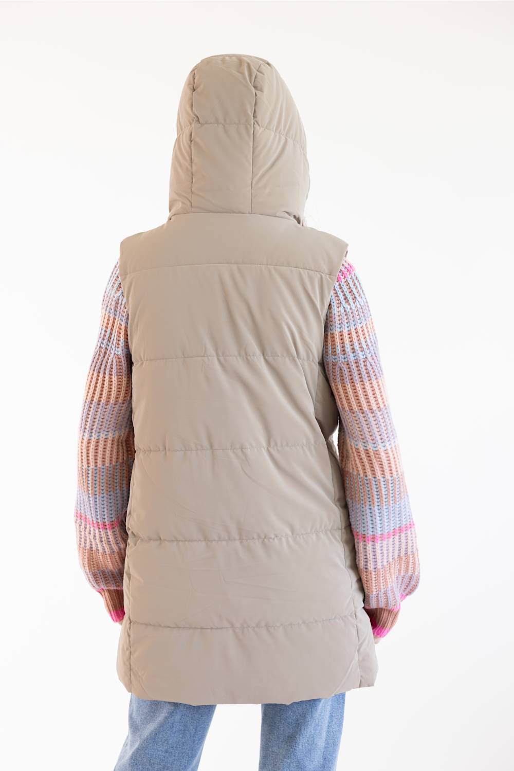 A person shown from the back wearing a light beige hooded puffer vest over a multicolored knit sweater and blue jeans, against a plain white studio background.