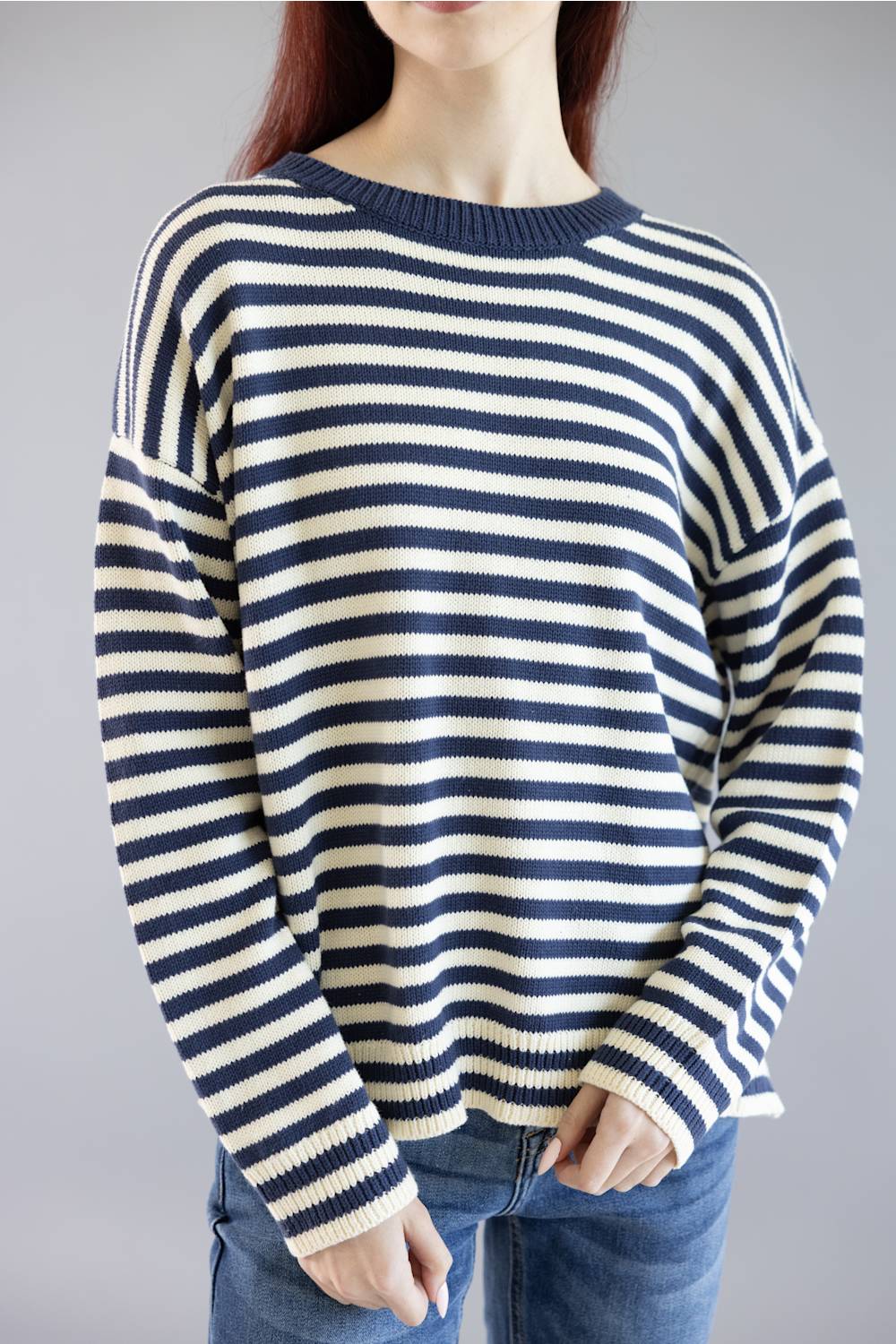 A woman wearing a navy blue and cream horizontal striped knit sweater with blue jeans, shown from the neck down against a plain gray studio background.