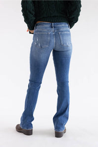 Rear view of a person wearing blue denim jeans with a dark green cable-knit sweater and dark brown shoes, standing against a plain white studio background.