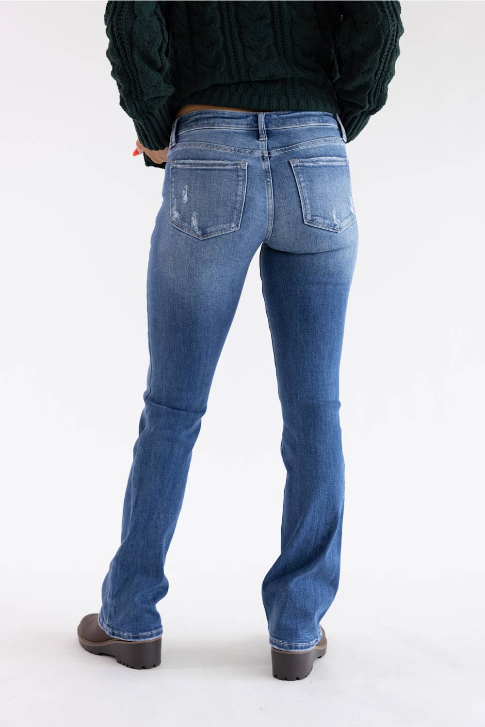 Rear view of a person wearing blue denim jeans with a dark green cable-knit sweater and dark brown shoes, standing against a plain white studio background.
