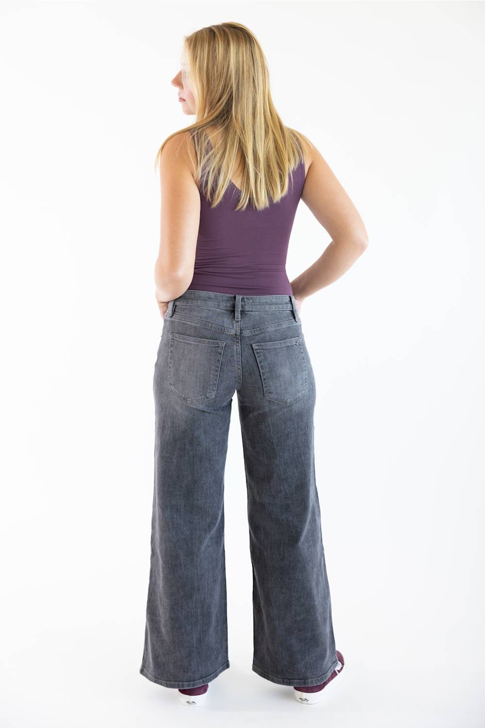 A woman shown from the back wearing wide-leg dark gray denim jeans and a purple sleeveless top, standing against a plain white studio background.