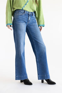 A model wearing a pair of medium-wash blue flare jeans, styled with a light green sweater and black heeled boots against a plain white studio background.