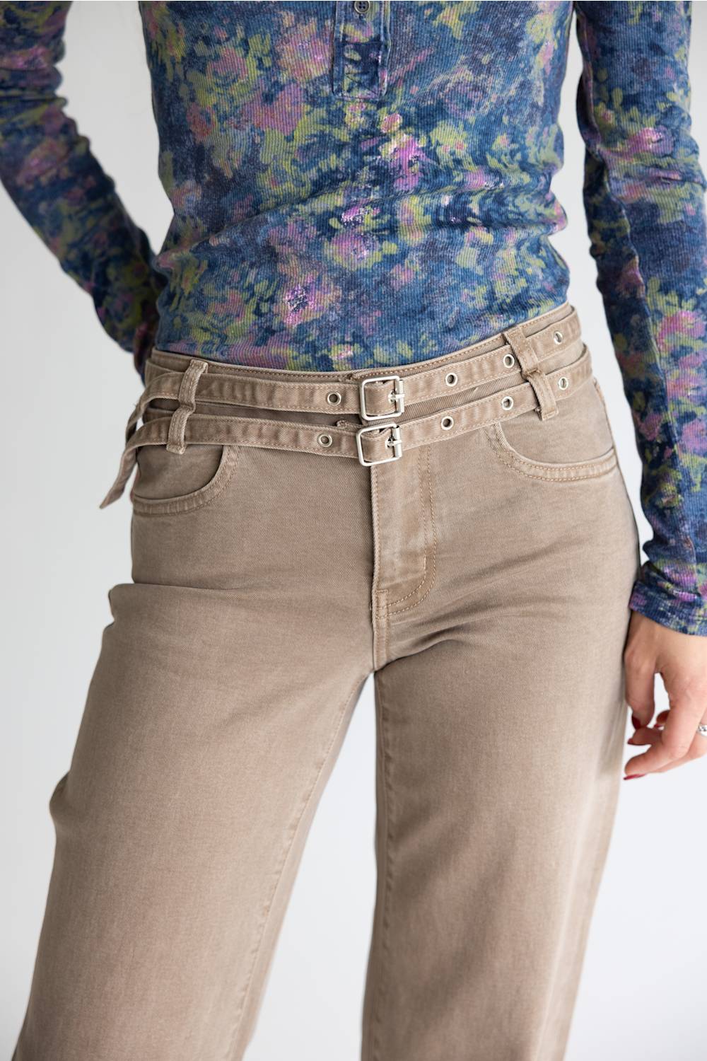 A close-up of a person wearing beige high-waisted jeans with a double-buckle belt and a blue floral long-sleeve top against a light gray background.
