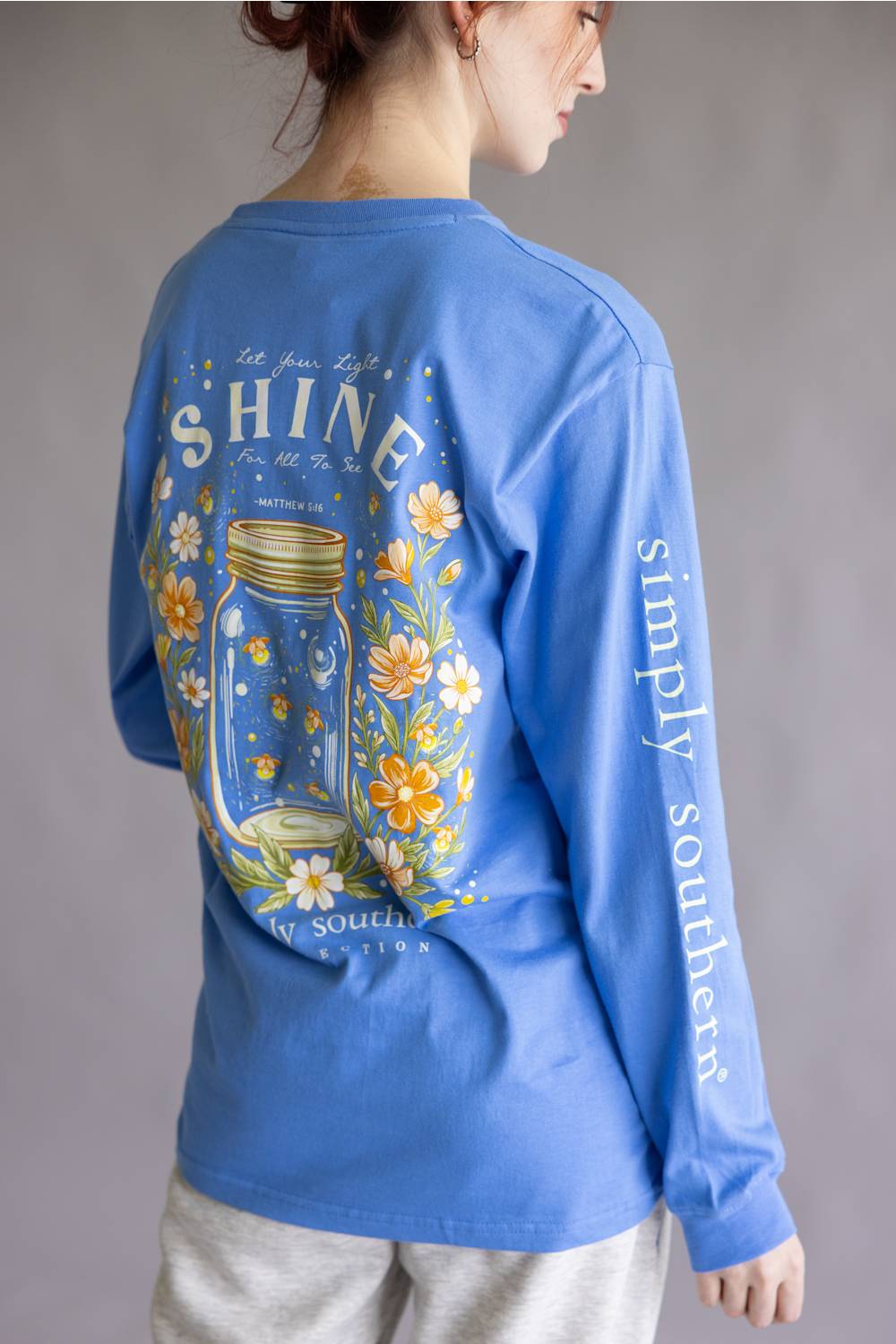 A woman shown from the back wearing a blue long-sleeve graphic T-shirt. The shirt features a large back print with a mason jar, yellow and white flowers, and the text 'SHINE' and 'simply southern', paired with light gray sweatpants against a neutral gray background.