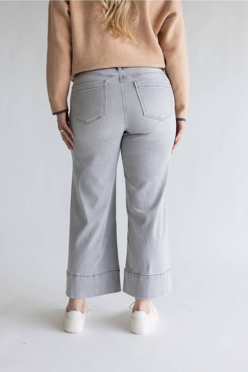A person shown from the back wearing light gray wide-leg cropped jeans, a beige sweater, and white sneakers against a plain light gray studio background.