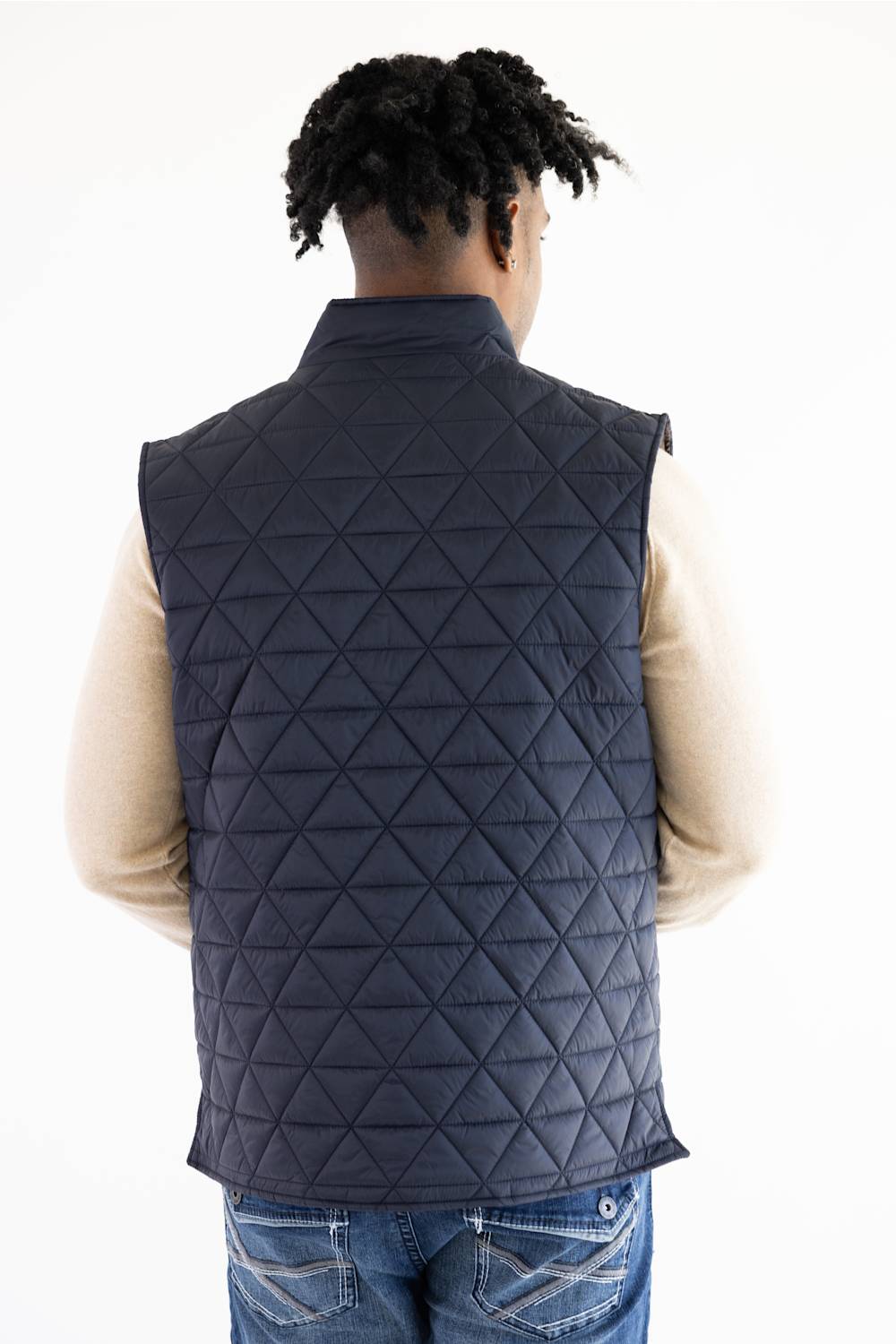 A man viewed from behind wearing a dark navy blue diamond-quilted puffer vest over a beige long-sleeve shirt and blue jeans, against a plain white background.