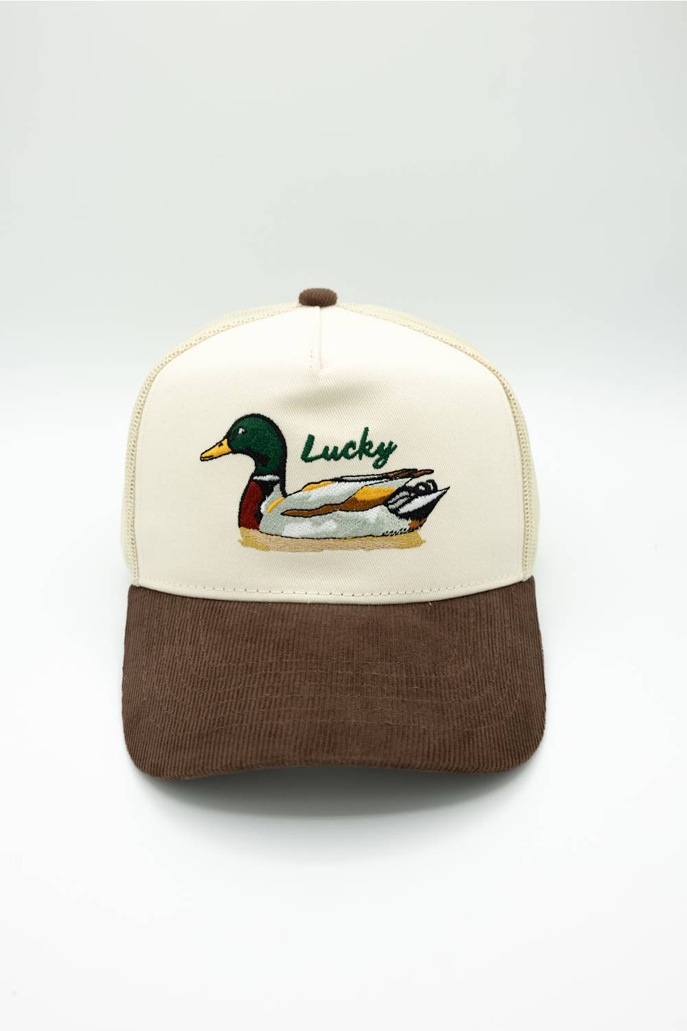 A trucker-style baseball cap with an off-white front panel and brim, a brown mesh back and curved brim, and an embroidered mallard duck with the word 'Lucky' on the front.