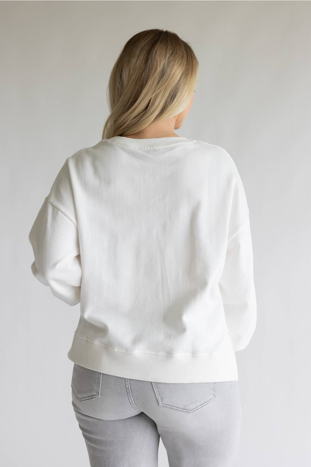 A woman viewed from behind wearing a plain white crewneck sweatshirt and light gray jeans, standing against a neutral light gray studio background.